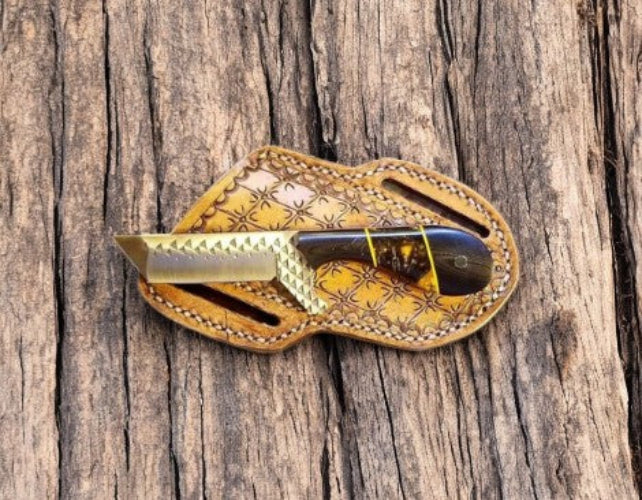 A folding pocket knife with a patterned tan leather sheath, resting on a rustic, textured wood surface.