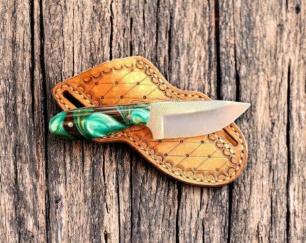 A fixed-blade knife with a green marbled handle and a silver blade, resting on a brown leather sheath with decorative stitching, placed on a rustic wooden surface.