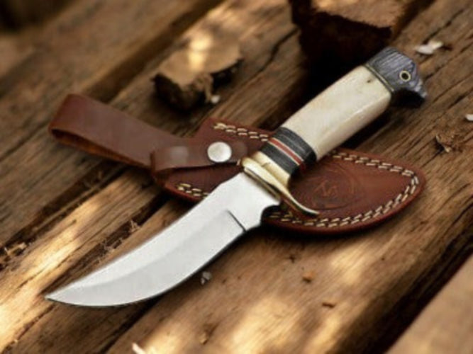 A fixed-blade knife with a curved silver blade, cream-colored handle, and brown leather sheath, resting on rustic wooden planks.