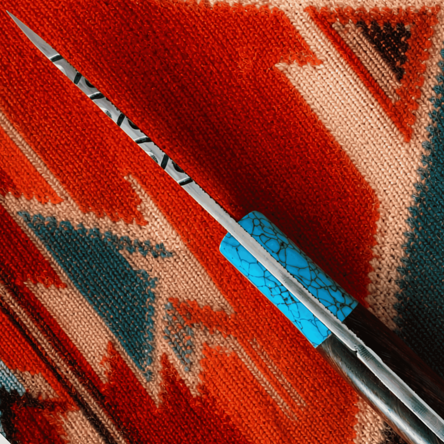 A close-up of a knife with a silver blade and a turquoise-patterned handle, resting diagonally on a red, orange, and beige woven textile.