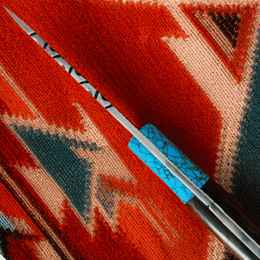 A close-up of a knife with a silver blade and a turquoise-patterned handle, resting diagonally on a red, orange, and beige woven textile.
