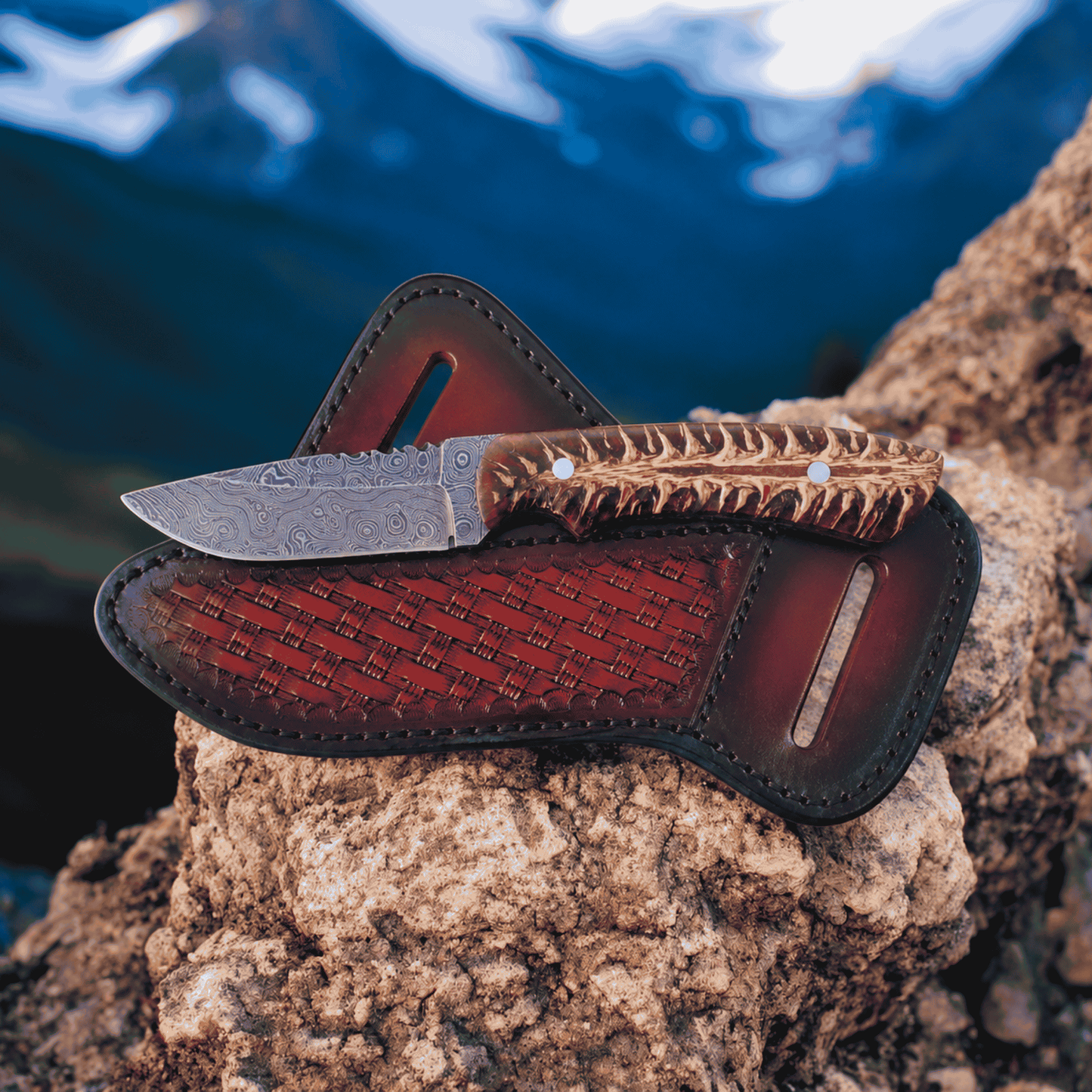 Bumpkin Ranchwear Rustic Damascus Cowboy Knife with pine cone resin handle set on a rock with mountains in the back ground.