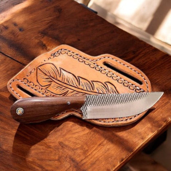 A fixed-blade knife with a dark wood handle and textured silver blade rests on a brown leather sheath with an embossed feather design, placed on a wooden table.