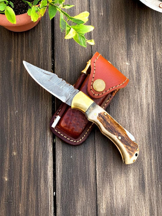 "Cowboy pocket knife with antler horn handle, Damascus blade, and brown leather sheath, laid on wooden surface."