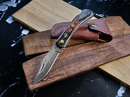 Folding knife with decorative blade and brown leather sheath on a wooden surface