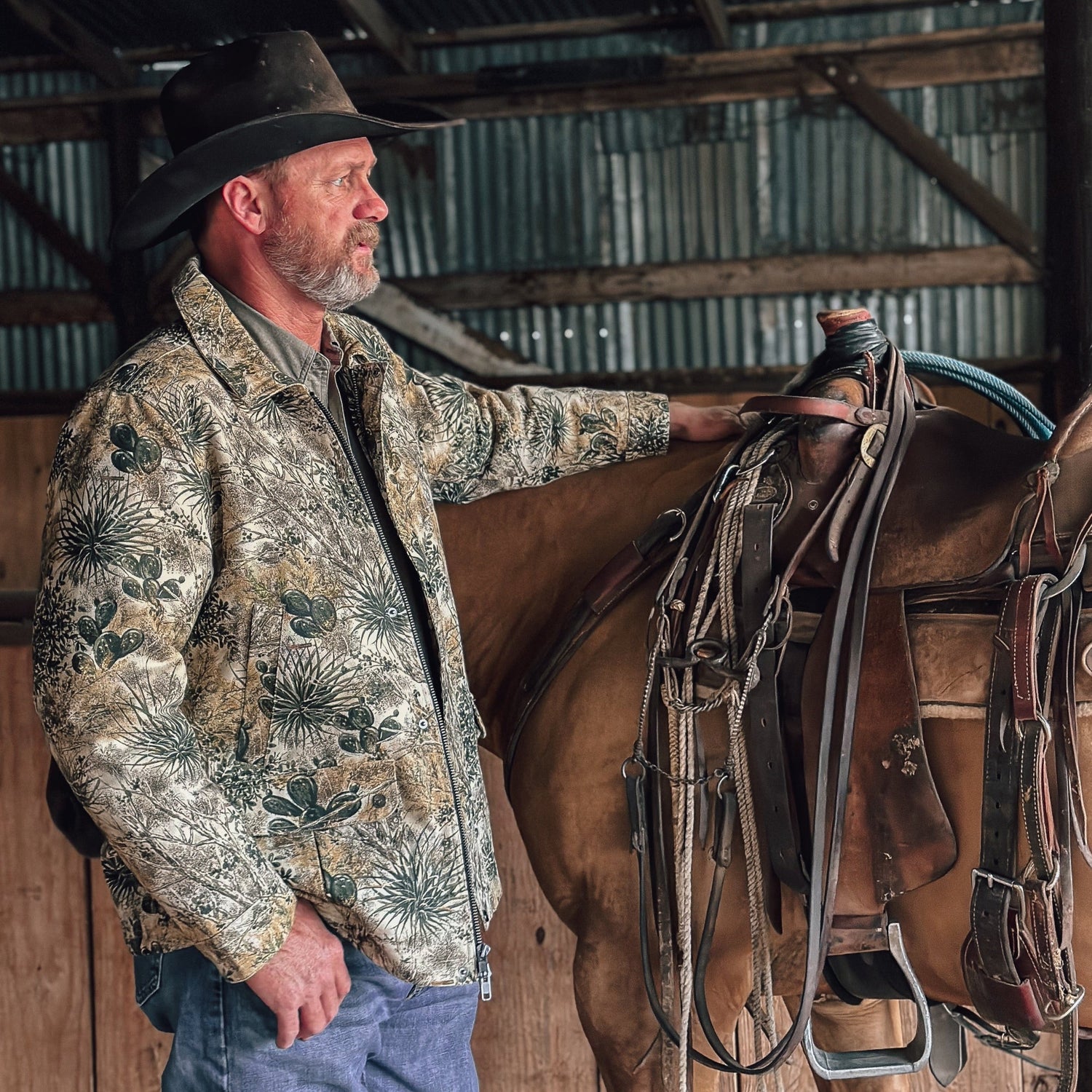 Man in a patterned jacket and cowboy hat standing next to a horse with a saddle in a barn.