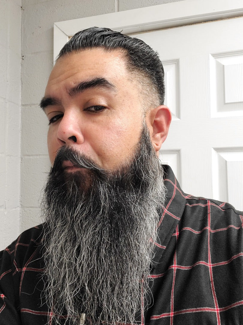 Man with a long gray beard and black hair wearing a plaid shirt indoors.