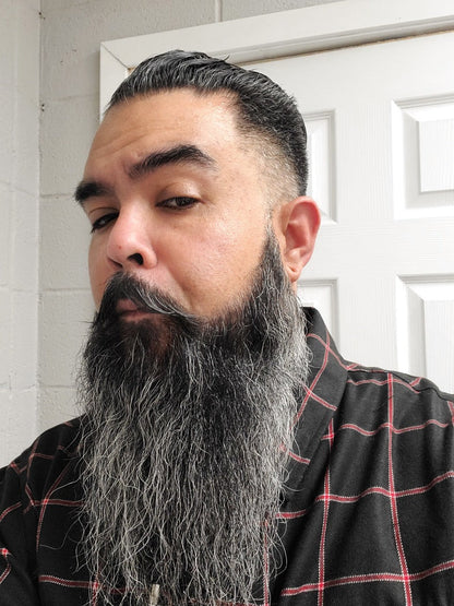 Man with a long gray beard and black hair wearing a plaid shirt indoors.