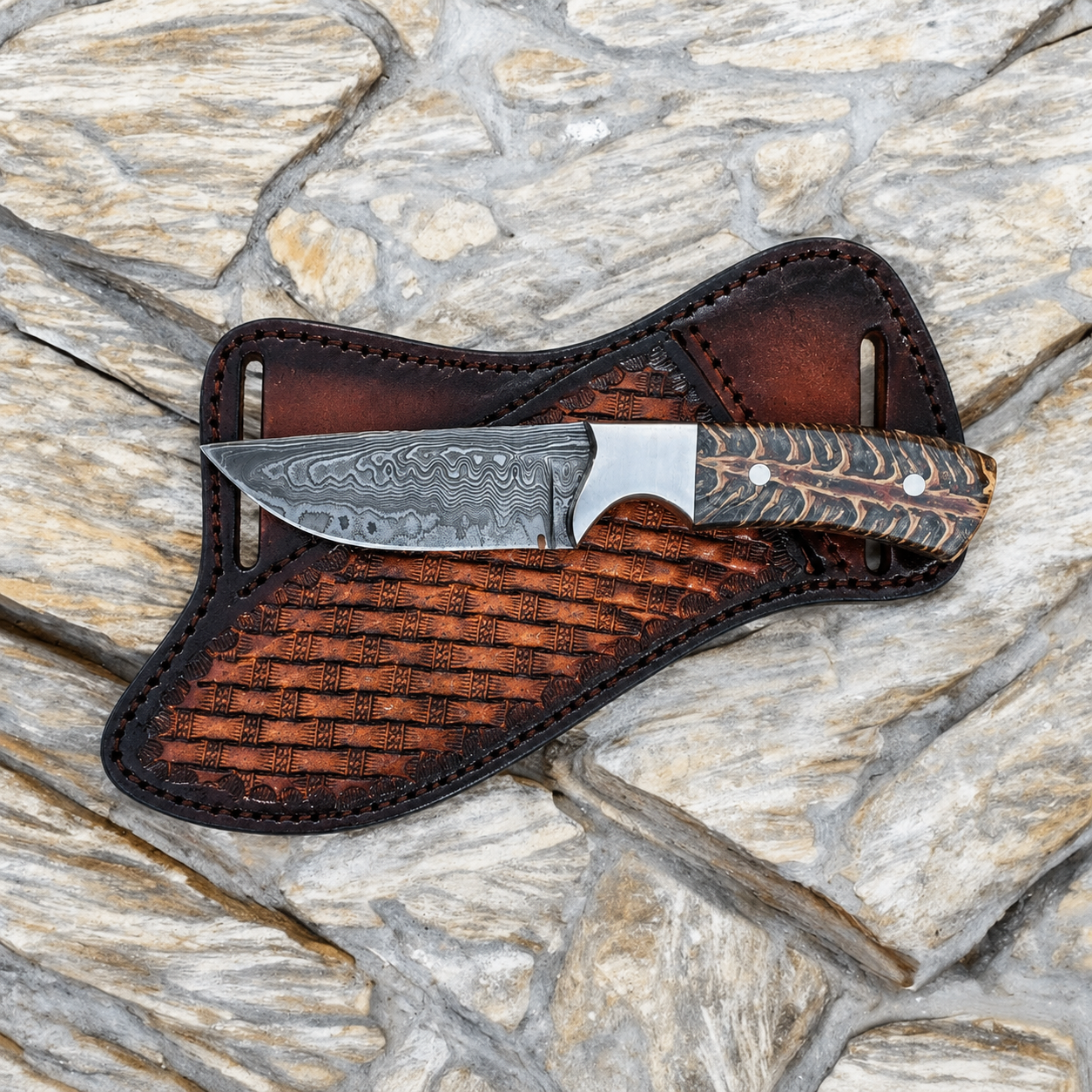 A fixed-blade knife with a patterned steel blade and a textured handle, resting on a dark brown leather sheath over a stone surface.