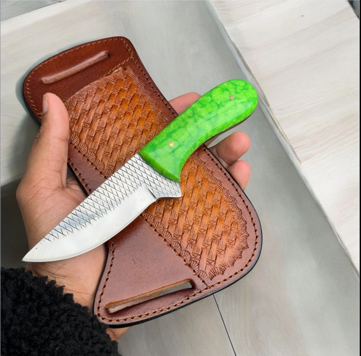 Hand holding a knife with a green handle and brown leather sheath.