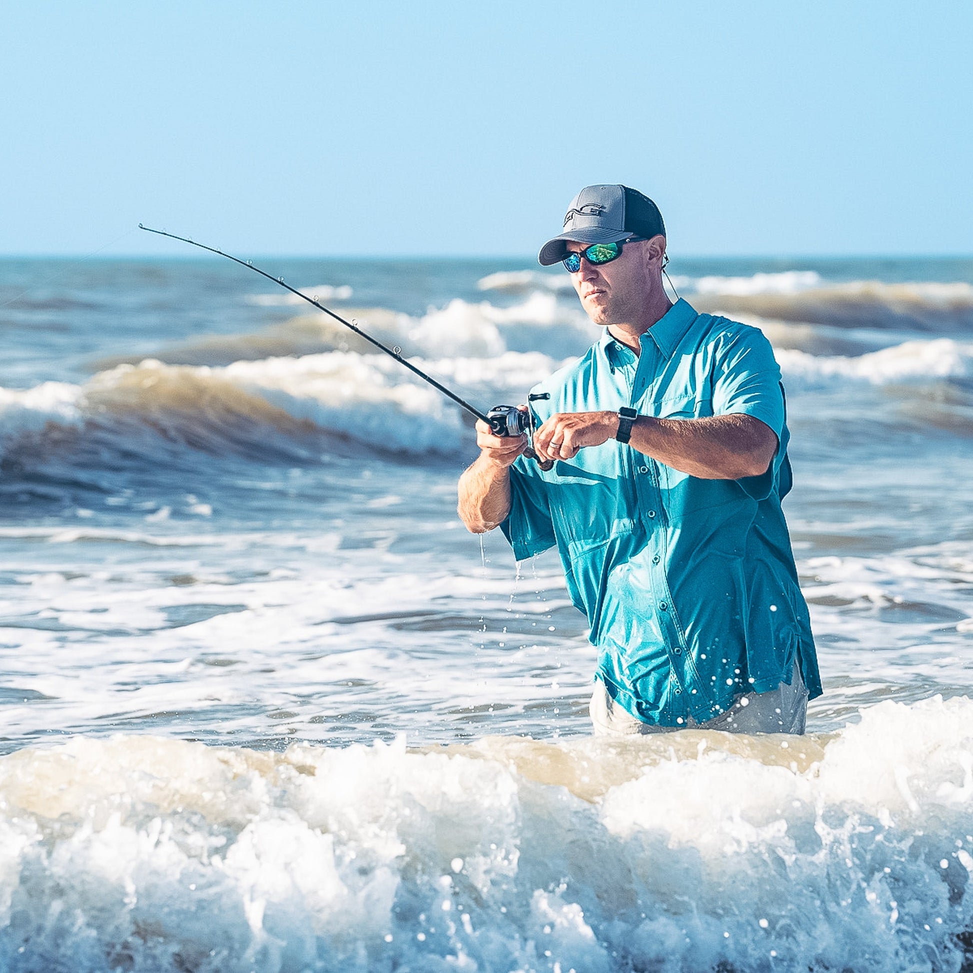 Man fishing in the ocean waves wearing a blue shirt and cap.