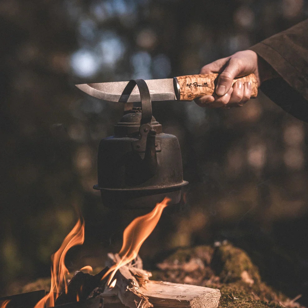 Person holding a knife over a small black pot over a campfire with a blurred natural background