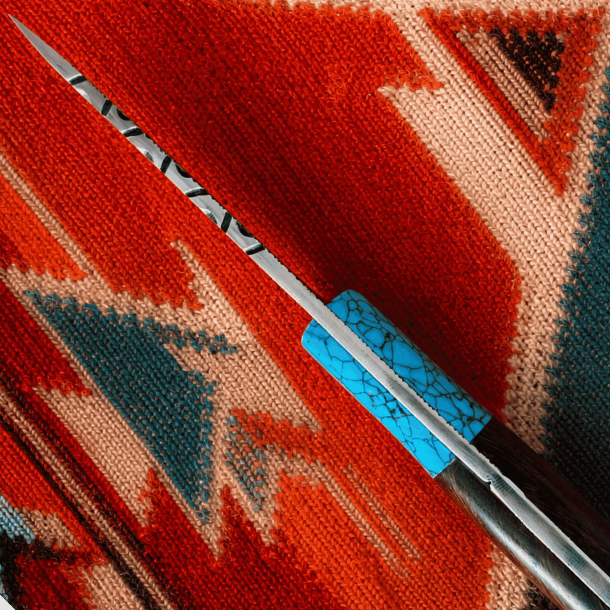 A close-up of a knife with a silver blade and a turquoise-patterned handle, resting diagonally on a red, orange, and beige woven textile.