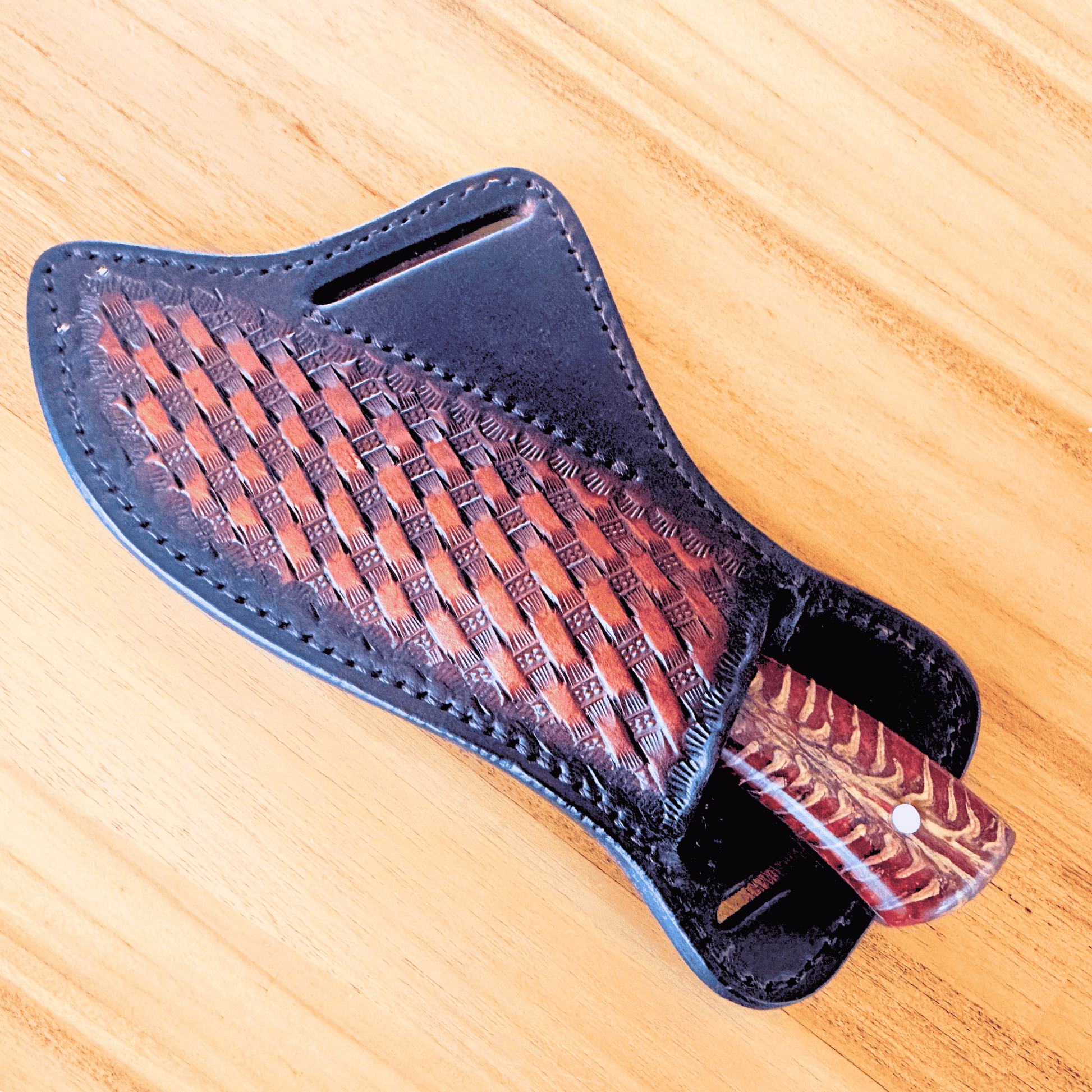 A handcrafted leather knife sheath with an embossed basketweave pattern, shown on a light wood grain background. The sheath is dark brown leather with a reddish-brown handle featuring a matching woven pattern.