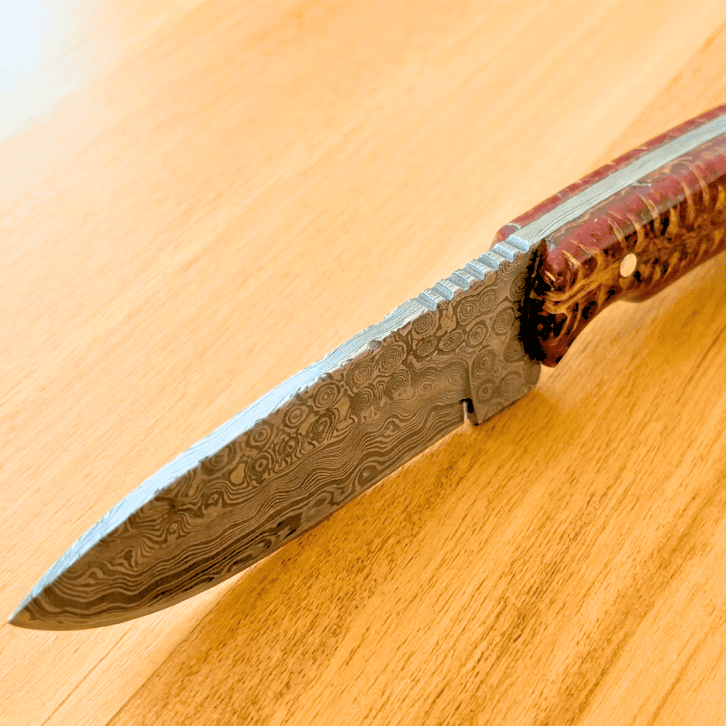A close-up of a Damascus steel folding knife with a patterned steel blade and a reddish-brown wood handle, resting diagonally on a light wood grain surface.