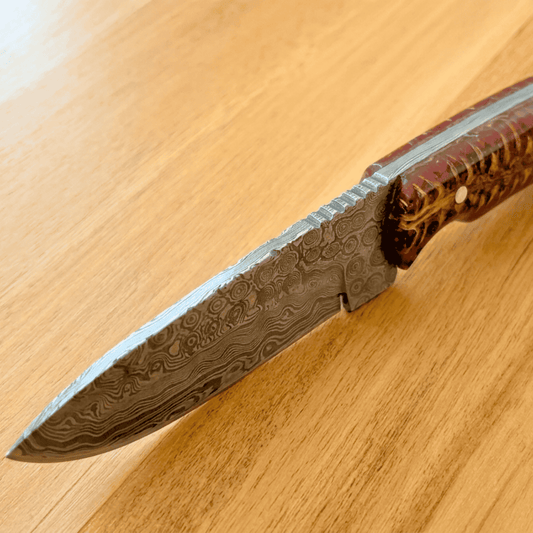 A close-up of a Damascus steel folding knife with a patterned steel blade and a reddish-brown wood handle, resting diagonally on a light wood grain surface.