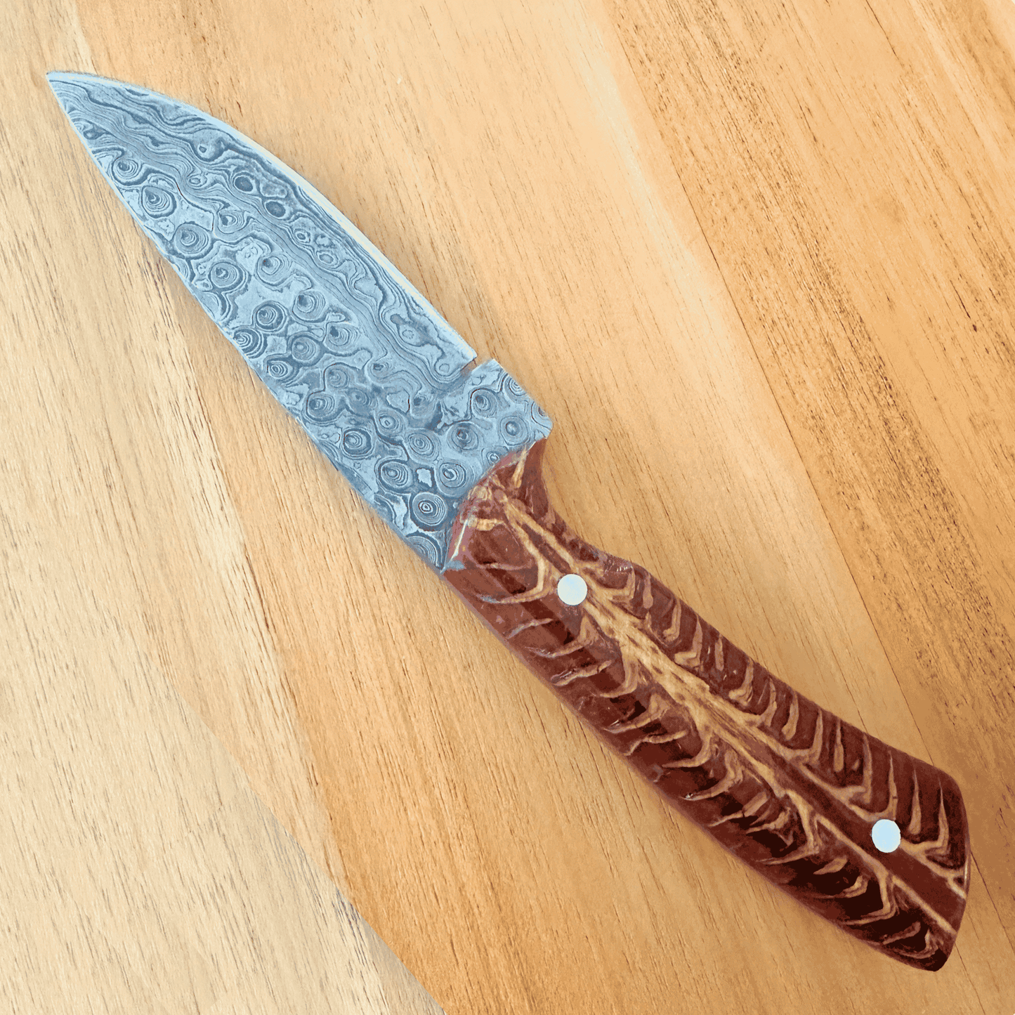A fixed-blade knife with a Damascus steel blade and a carved red brown handle, placed diagonally on a light wood surface.