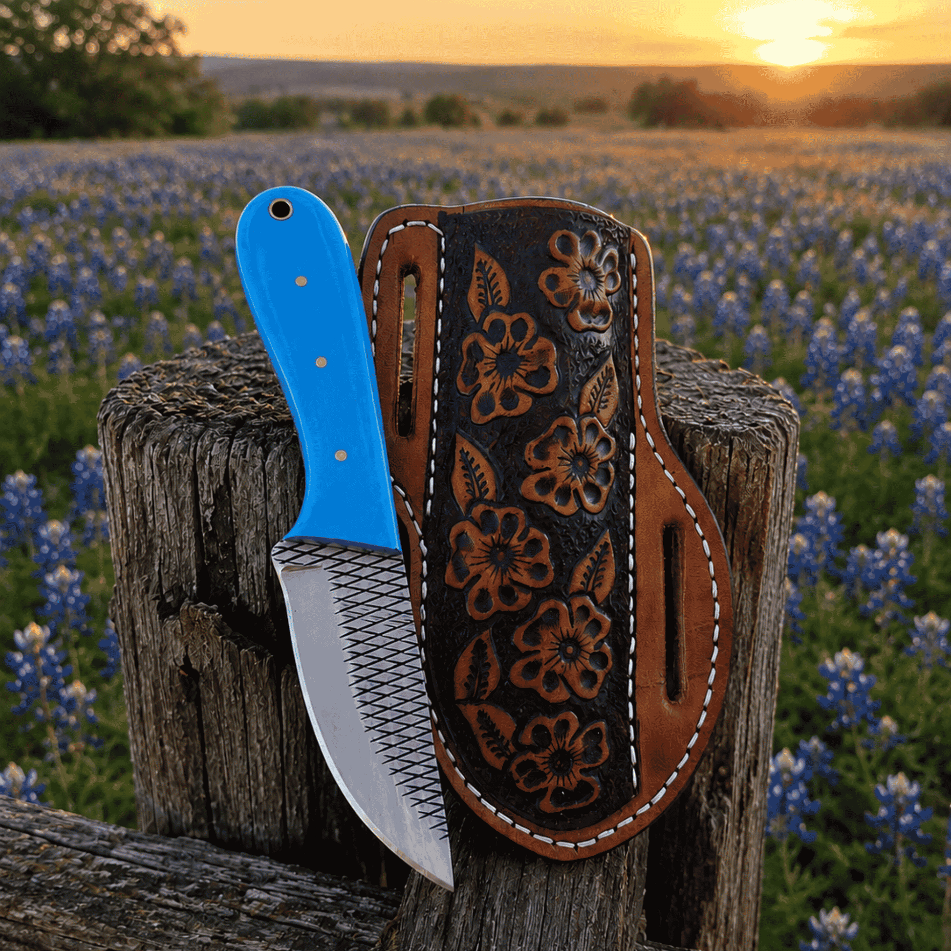 A custom knife with a bright blue handle and patterned steel blade is displayed next to its hand-tooled brown leather sheath on a rustic wooden fence post, with a field of bluebonnets and a sunset in the background.