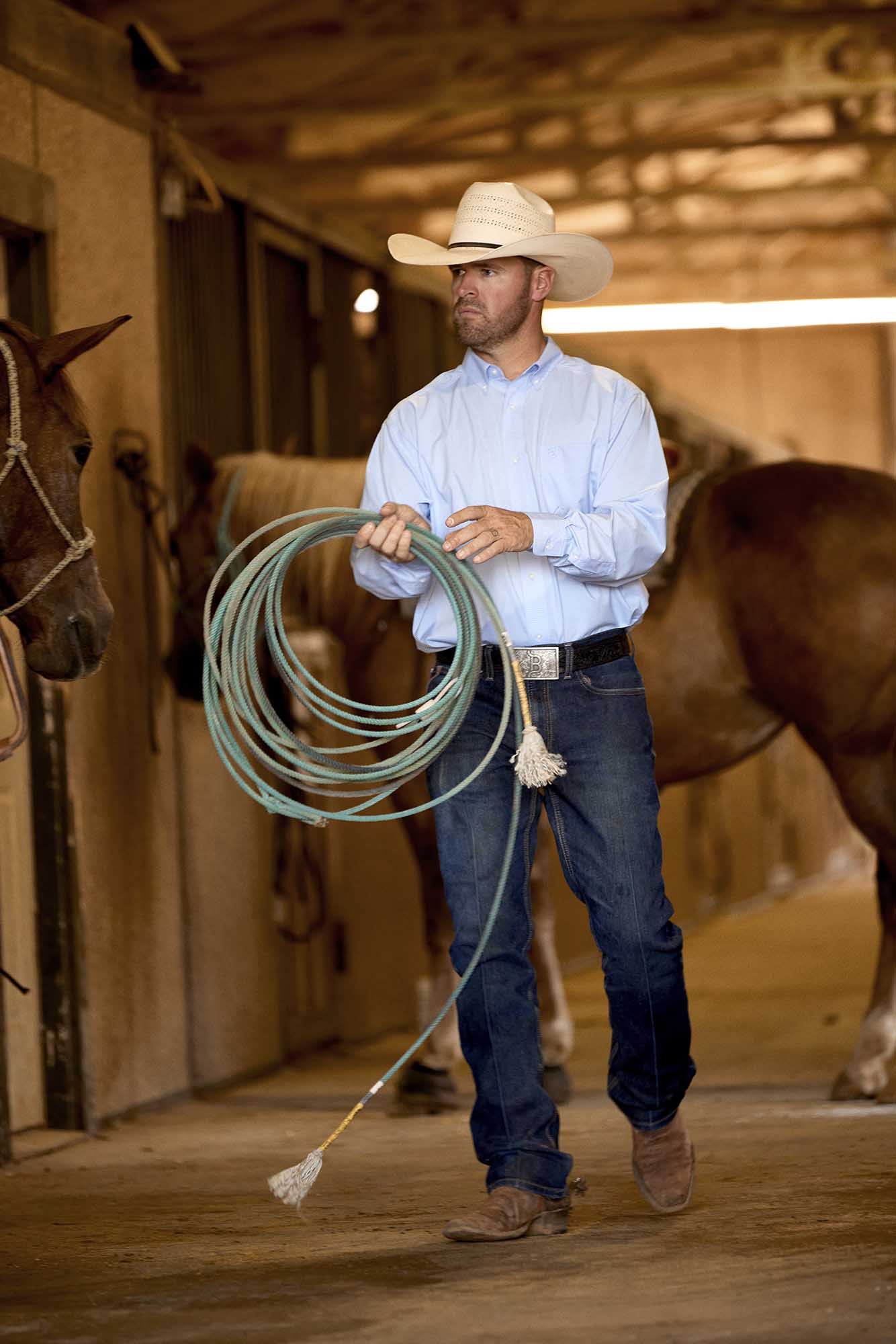Man holding a lasso in a stable with horses in the background wearing Ariat button down men's western shirt