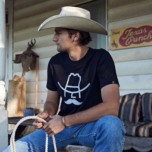 Man wearing a cowboy hat and black t-shirt with a white graphic, sitting on a porch.